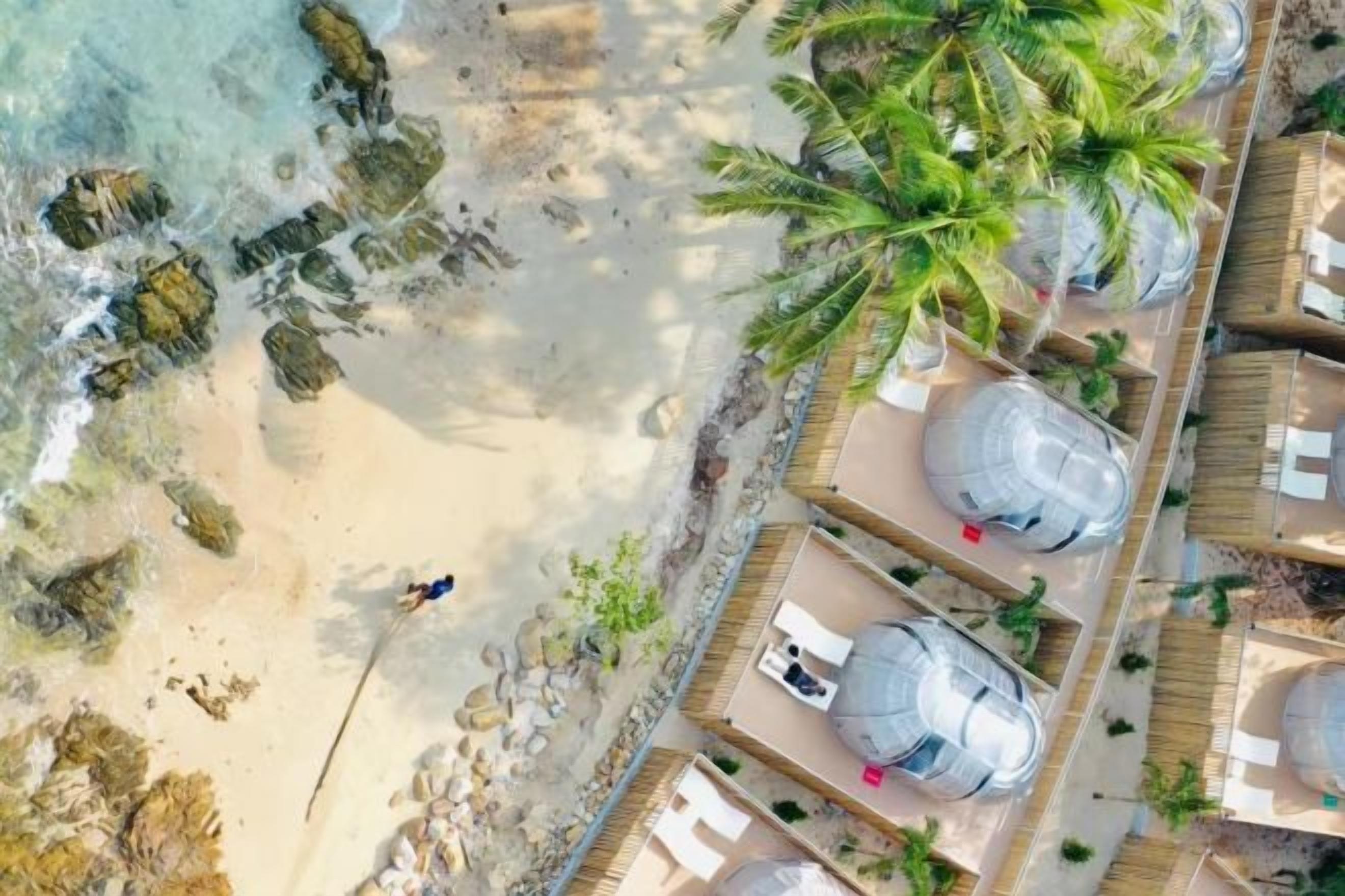 A beachside resort with palm trees, a sandy beach, and several large cylindrical structures, possibly water tanks.