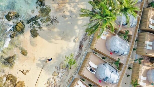 Vue aérienne d’un complexe hôtelier comprenant plusieurs structures sphériques, une plage de sable et des palmiers.