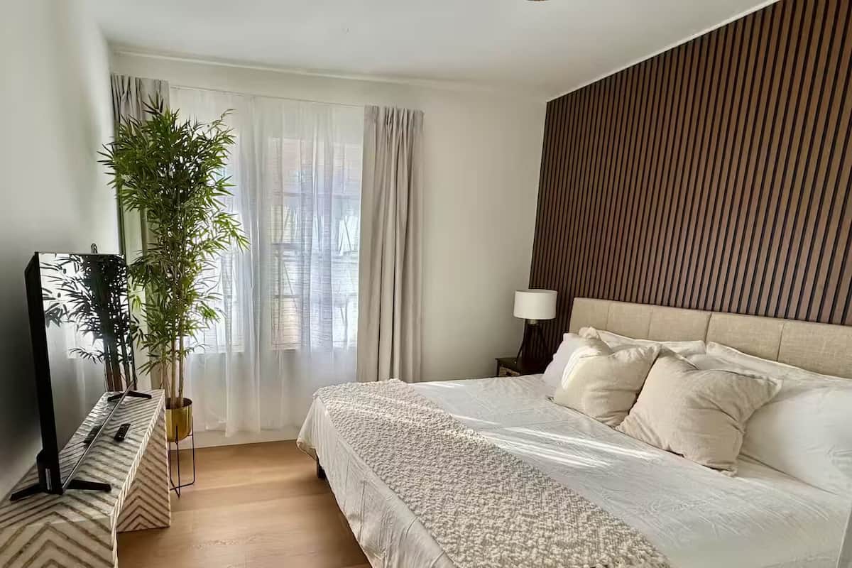 Une chambre d’hôtel moderne avec un grand lit, un téléviseur à écran plat, une plante en pot et une tête de lit en bois.