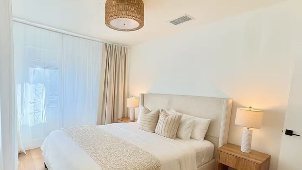Une chambre à coucher bien rangée, avec un grand lit, deux tables de chevet avec des lampes, une fenêtre avec des rideaux et un luminaire au plafond.