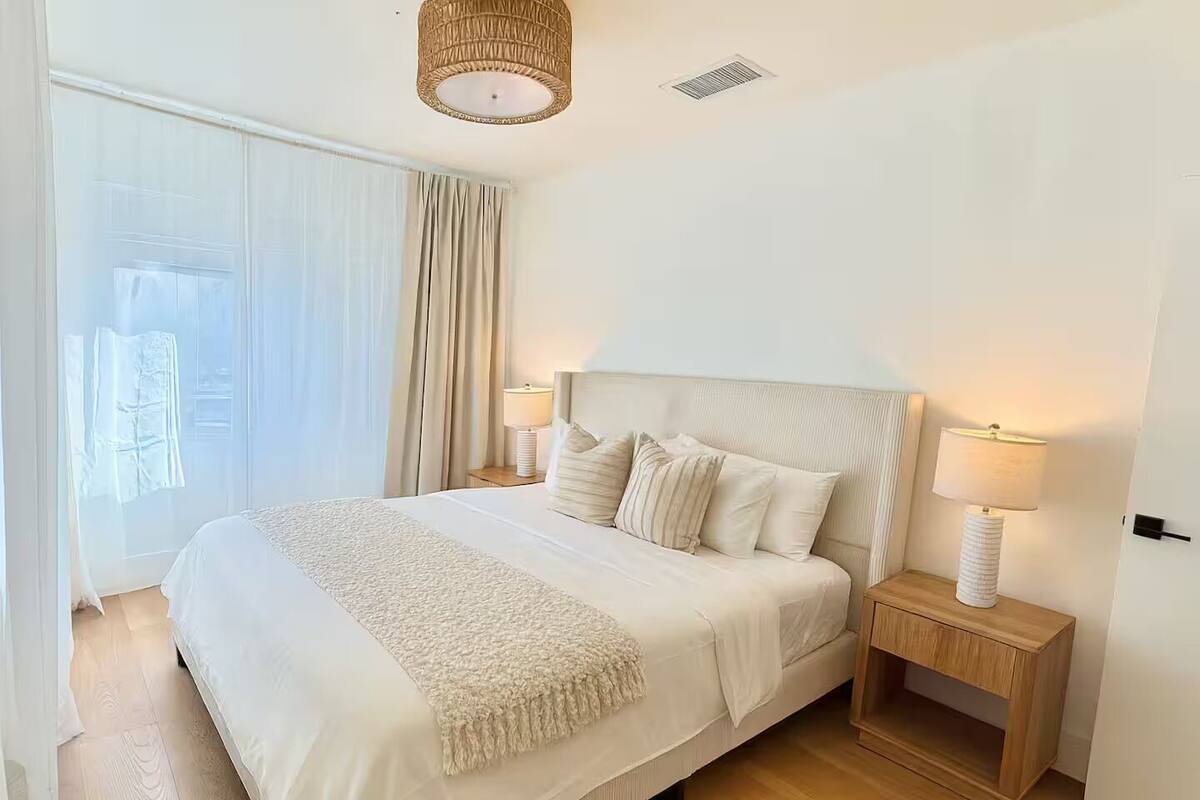 Une chambre à coucher bien rangée, avec un grand lit, deux tables de chevet avec des lampes, une fenêtre avec des rideaux et un luminaire au plafond.