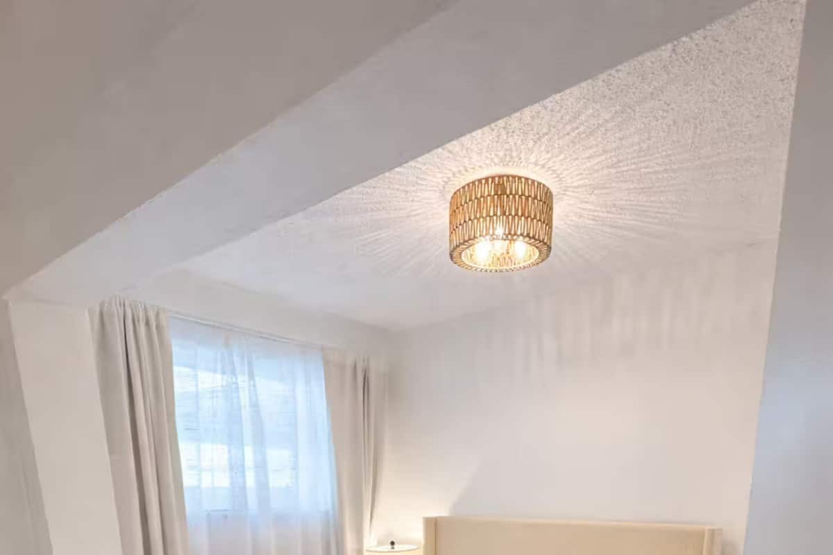 Un lit bien fait, avec du linge de lit blanc et un plaid beige, une table de chevet avec une lampe et un luminaire au plafond.
