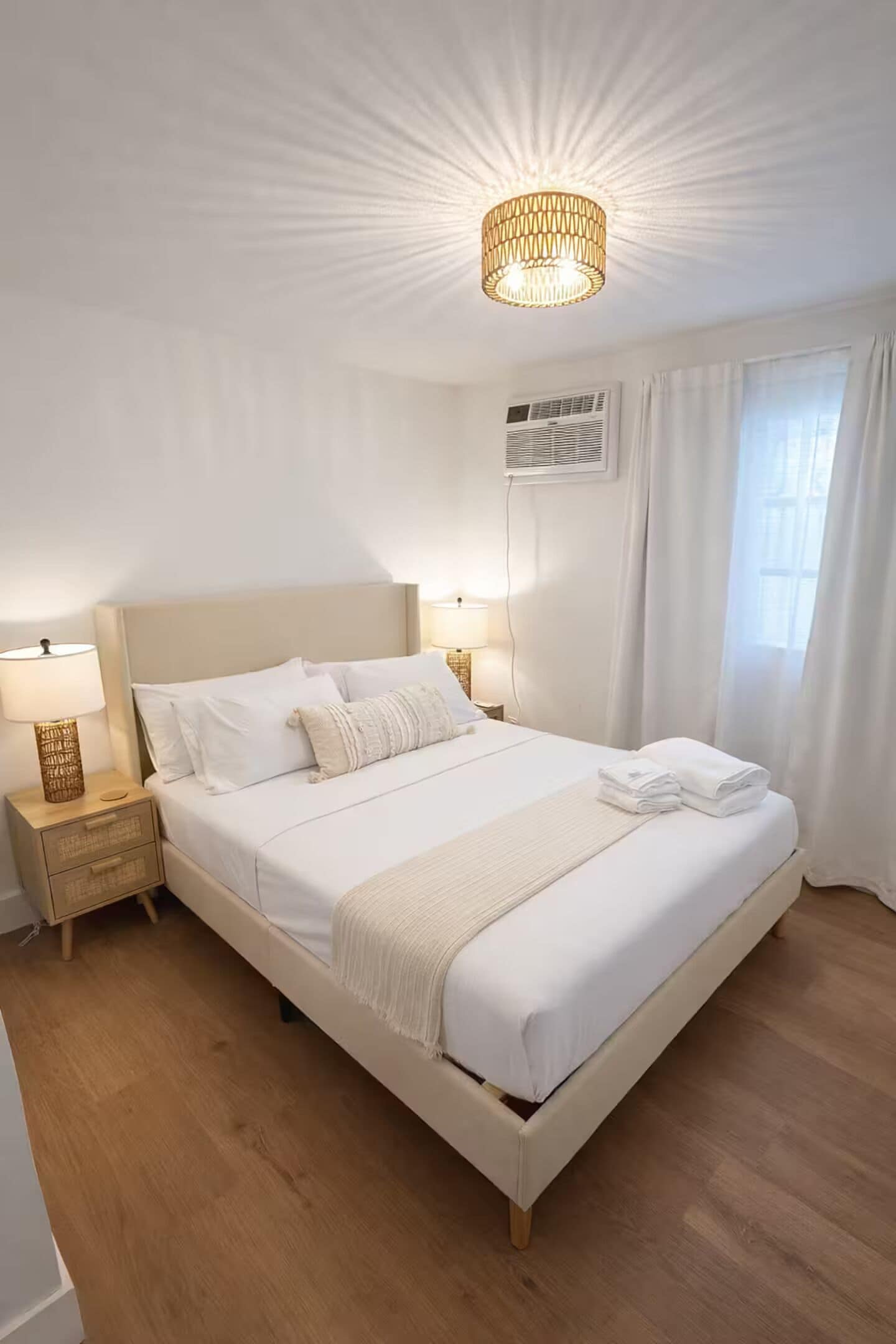 A neatly arranged bedroom with a large bed, bedside lamps, a ceiling light, and a window with curtains.