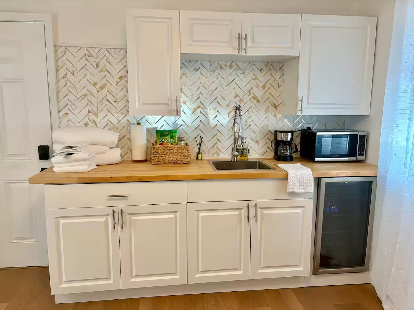 Cocina compacta con armarios blancos, un salpicadero con estampado de espiga, microondas, cafetera y refrigerador pequeño.
