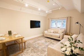 A living room with a sofa, a dining table set with plates and glasses, a TV mounted on the wall, and a window with curtains. - Brand New Modern 2-Bed Apartment | Broadway High St | Sleeps 4 (Worcestershire)