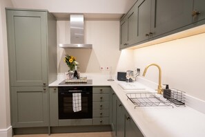 A kitchen with a built-in oven, a stainless steel range hood, a sink with a gold faucet, and a wire basket on the countertop. - Brand New Modern 2-Bed Apartment | Broadway High St | Sleeps 4 (Worcestershire)