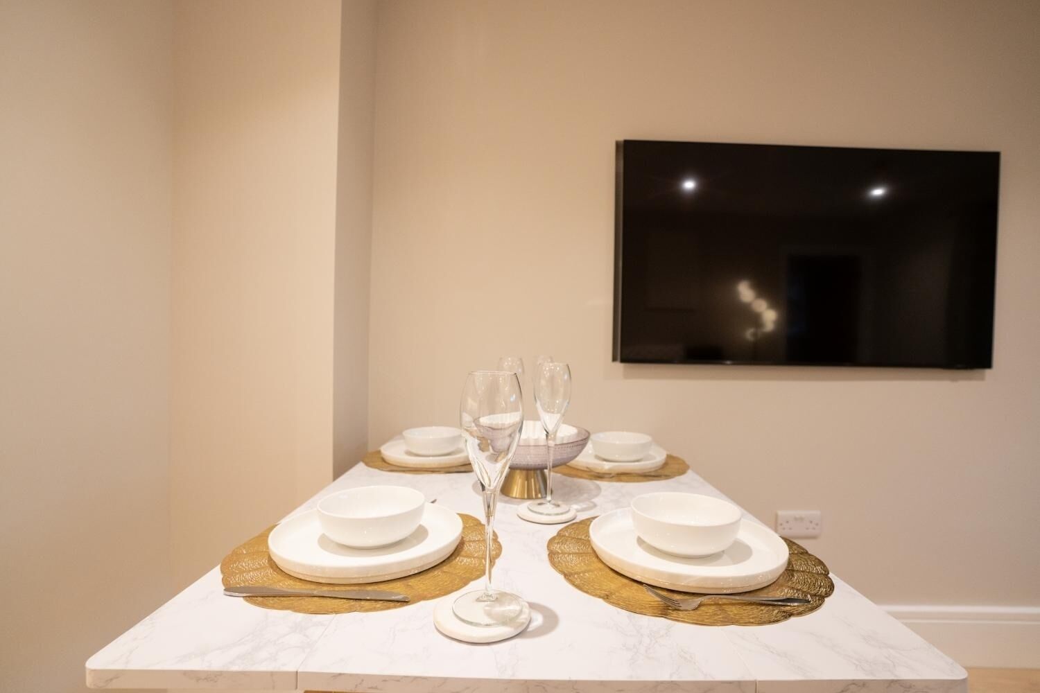 A dining table set with white plates, bowls, and glasses, with a gold placemat and a framed picture on the wall behind.