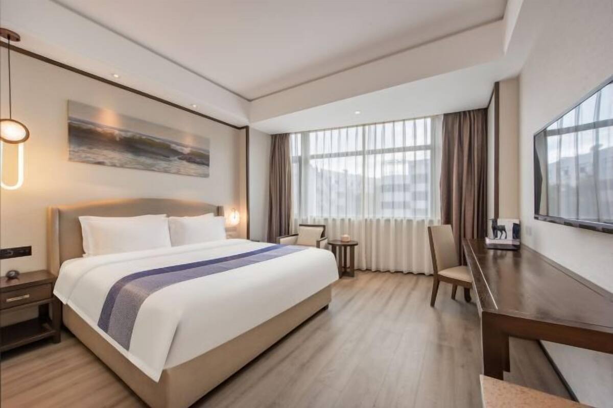 Ett hotellrum med en stor säng, ett skrivbord, en stol, ett litet bord och en tavla på väggen.
