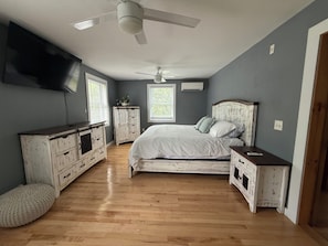 A bedroom with a bed, dresser, nightstand, ceiling fan, and a TV mounted on the wall.