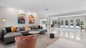 A modern living room with a grey sofa, a coffee table, and a pink chair. There are two large paintings on the wall, a lamp, and a dining area with a glass table and chairs. - Beautiful 2 bedroom bungalow in a quiet neighborhood (Fort Myers)