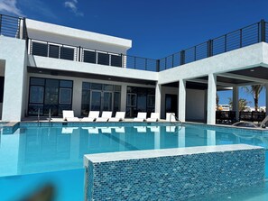 Uma área de piscina moderna com uma piscina azul cristalina, cadeiras de descanso brancas e um edifício de dois andares com janelas amplas e uma varanda.