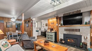 A cabin interior with a fireplace, wooden furniture, a dining area, and a kitchen. - Mt Rainier Cabin on Big Creek! Hot Tub! Fireplace! (Ashford)