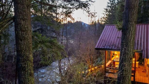 A cabin with a wooden deck and a small porch, surrounded by dense forest and a river. - Mt Rainier Cabin on Big Creek! Hot Tub! Fireplace! (Ashford)