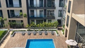A rooftop swimming pool with a wooden deck, surrounded by lounge chairs and umbrellas, and a modern apartment building in the background. - Esthetique Homes Embassy Gardens (Accra)