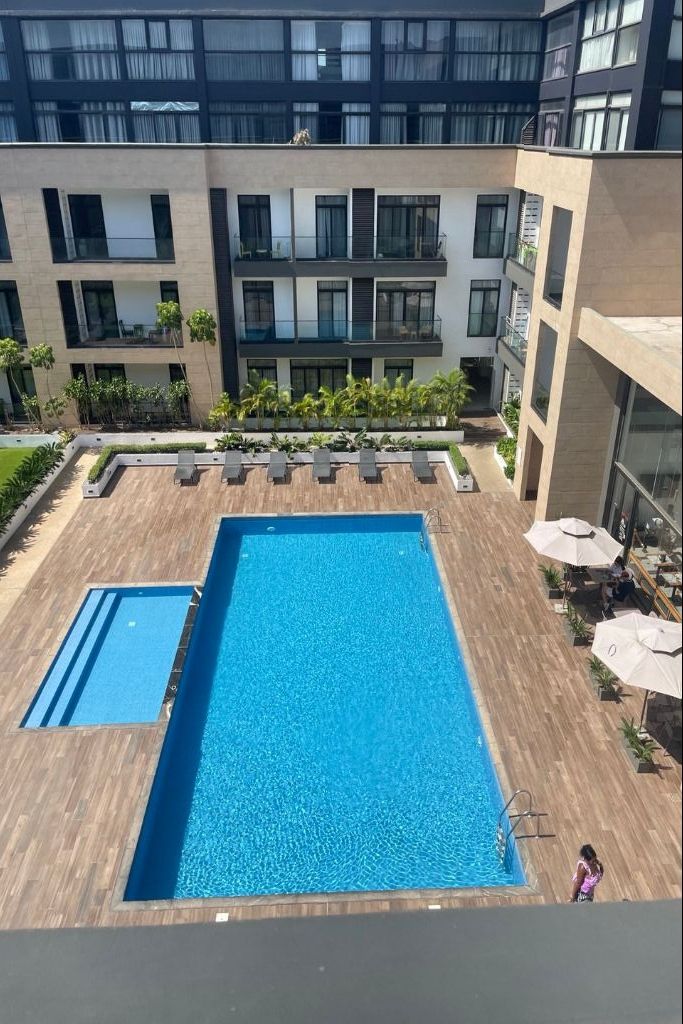 A rooftop pool with a wooden deck, surrounded by lounge chairs and umbrellas, with a modern building in the background.