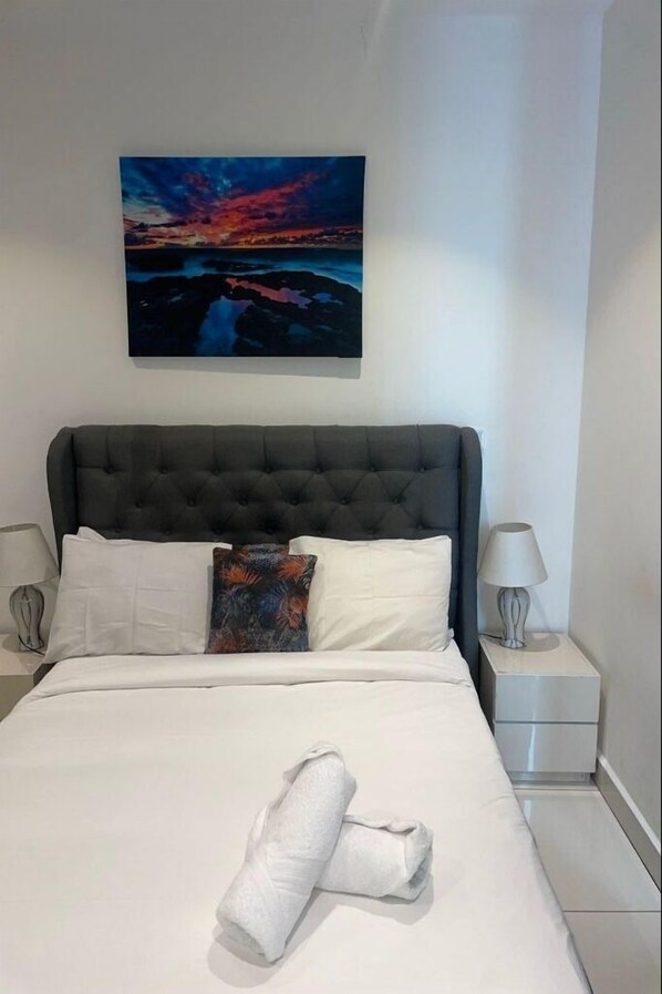 A neatly made bed with white bedding and a rolled towel, a dark tufted headboard, a colorful abstract painting above the bed, and two bedside tables with lamps. - Esthetique Homes Embassy Gardens (Accra)