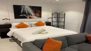 A modern hotel room with a large bed, two orange pillows, a grey sofa, a white towel, and a painting on the wall. - Esthetique Homes Embassy Gardens (Accra)
