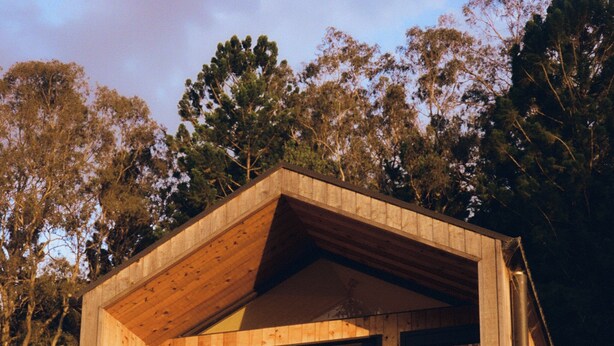 Una cabaña de madera con techo inclinado, ventanas grandes y un pequeño patio con sillas.
