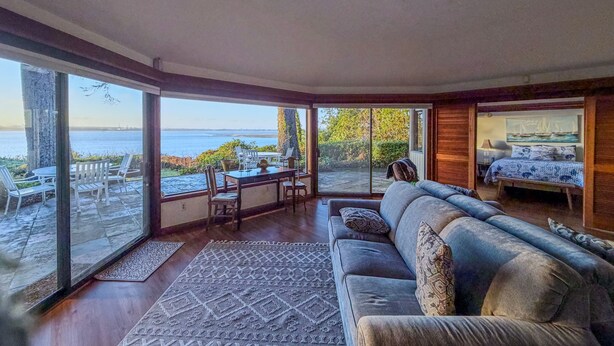 A modern living room with a large sliding glass door leading to an outdoor patio. Inside, there is a grey sofa, a dining table with chairs, and a small bed with a view of the sea. - The Seagates Private Coastal Retreat | Coos Bay (Coos Bay)