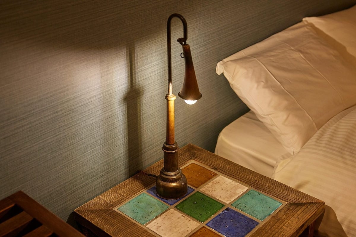 A bedside table with a vintage-style lamp, a patterned tablecloth, and a bed with white linens.