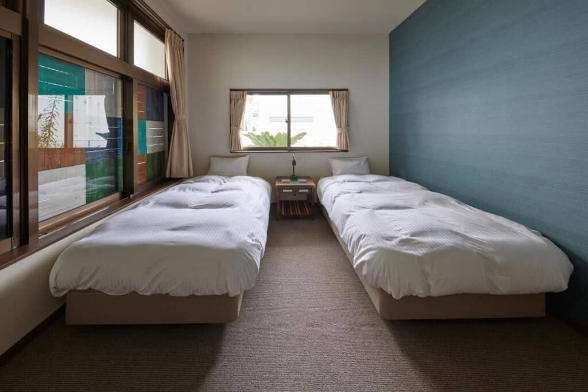 A hotel room with two single beds, a small table with a lamp, and a window with curtains.