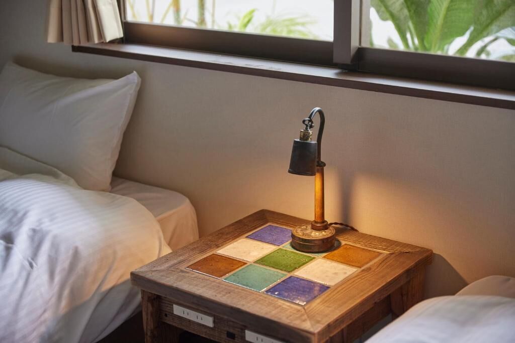 Une table de chevet en bois, une lampe, un lit et une fenêtre avec des rideaux.