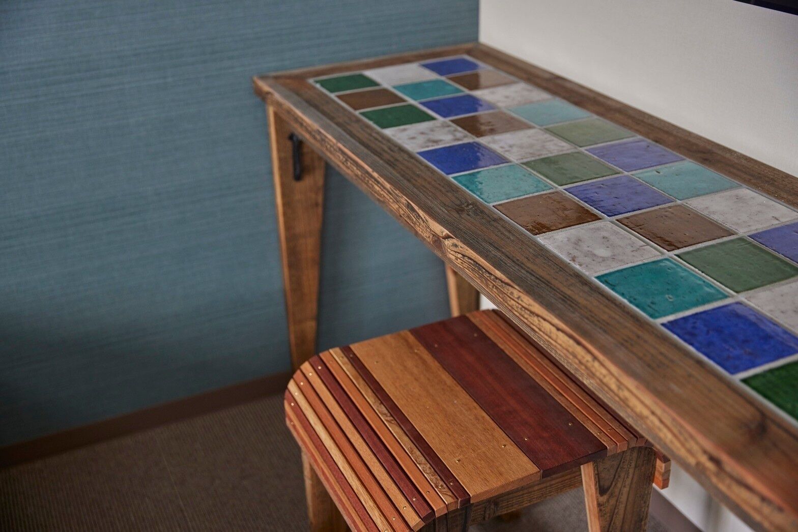 A wooden table with a colorful tile top, a matching wooden stool, and a blue wall in the background.