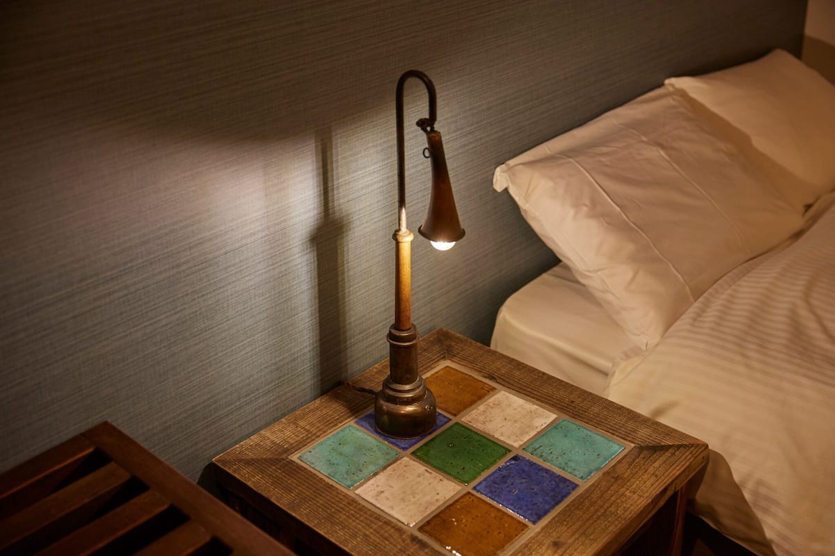 A bedside table with a vintage-style lamp, a patterned tray, and a bed with white bedding.