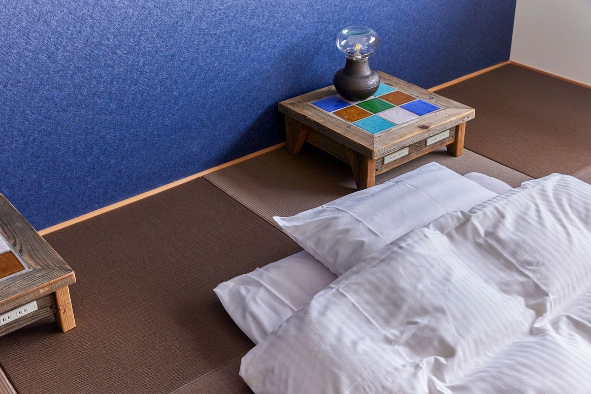 A traditional Japanese-style room with tatami flooring, a small wooden table with a lamp, and a bed with white bedding.