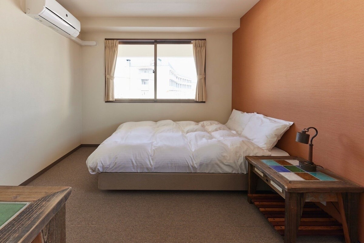 A hotel room with a bed, a window with curtains, a bedside table with a lamp, and a view of buildings outside.