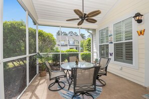 Eine überdachte Terrasse mit einem Glastisch und Stühlen, einem Deckenventilator und Blick auf Häuser im Hintergrund.