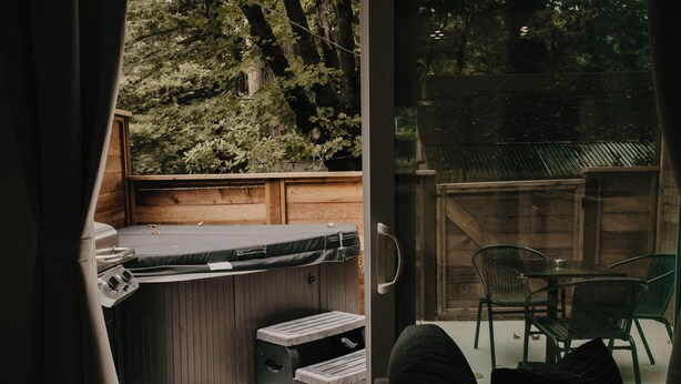 A hot tub with a wooden deck and a glass door leading to a patio area with a table and chairs. - Parkway Grove Cabins 1 Bedroom with Private Hot Tub and Spa Shower (Miranda)