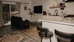 A modern kitchen with a white countertop, a coffee machine, and a sink. There is a black sofa and a brown chair in the living area. - Parkway Grove Cabins 1 Bedroom with Private Hot Tub and Spa Shower (Miranda)
