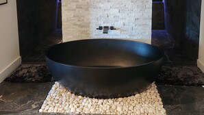 A modern bathroom with a large black bathtub, a stone wall, and a tiled floor. - Luxury Lake Norman Home (Mooresville)