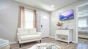 Un salon avec un canapé blanc, une table basse ronde avec un plateau en verre, un téléviseur fixé au mur et une porte avec une fenêtre.