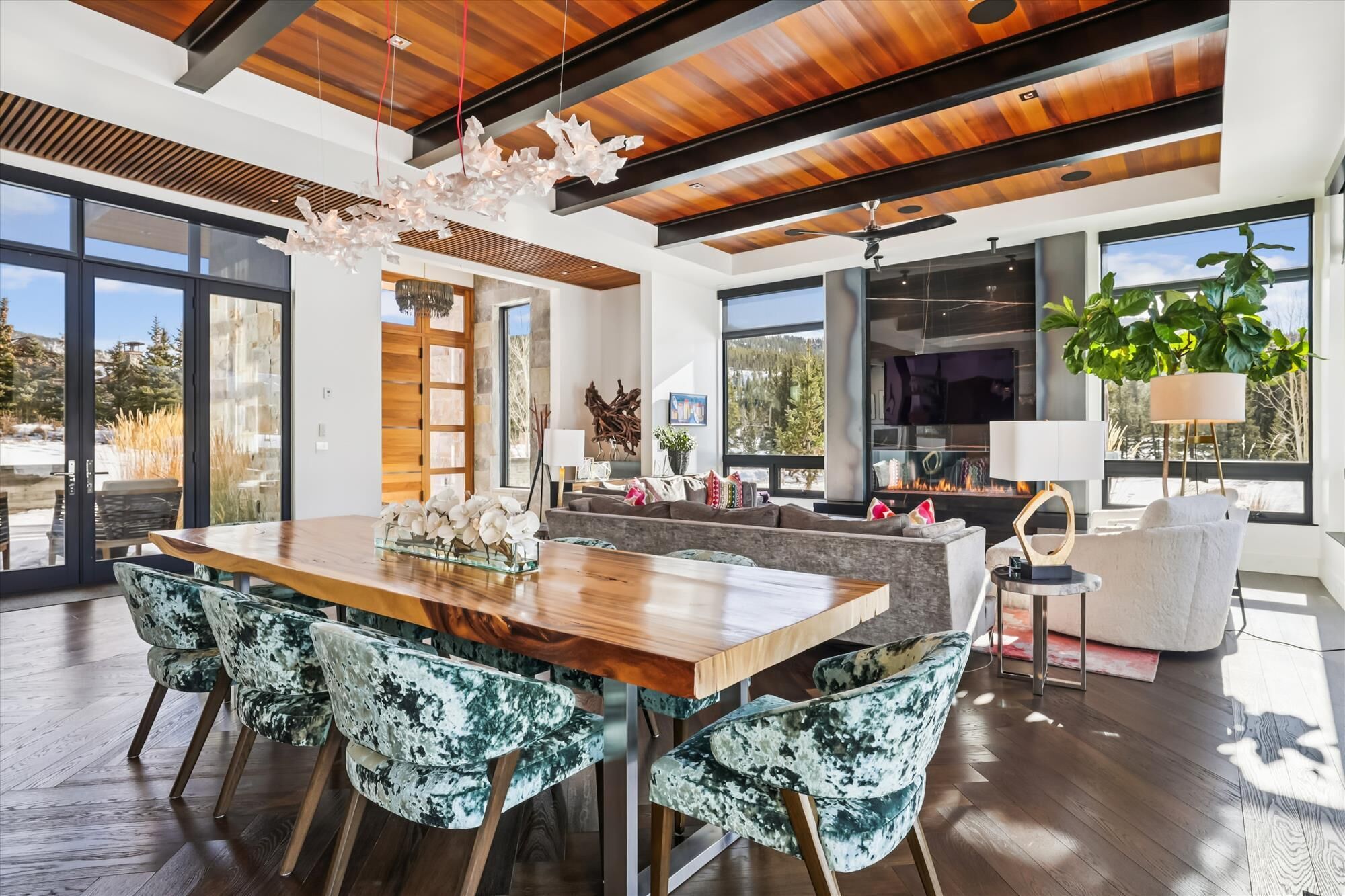 A modern living room with a wooden dining table, patterned chairs, a fireplace, and large windows offering a view of the outdoors.