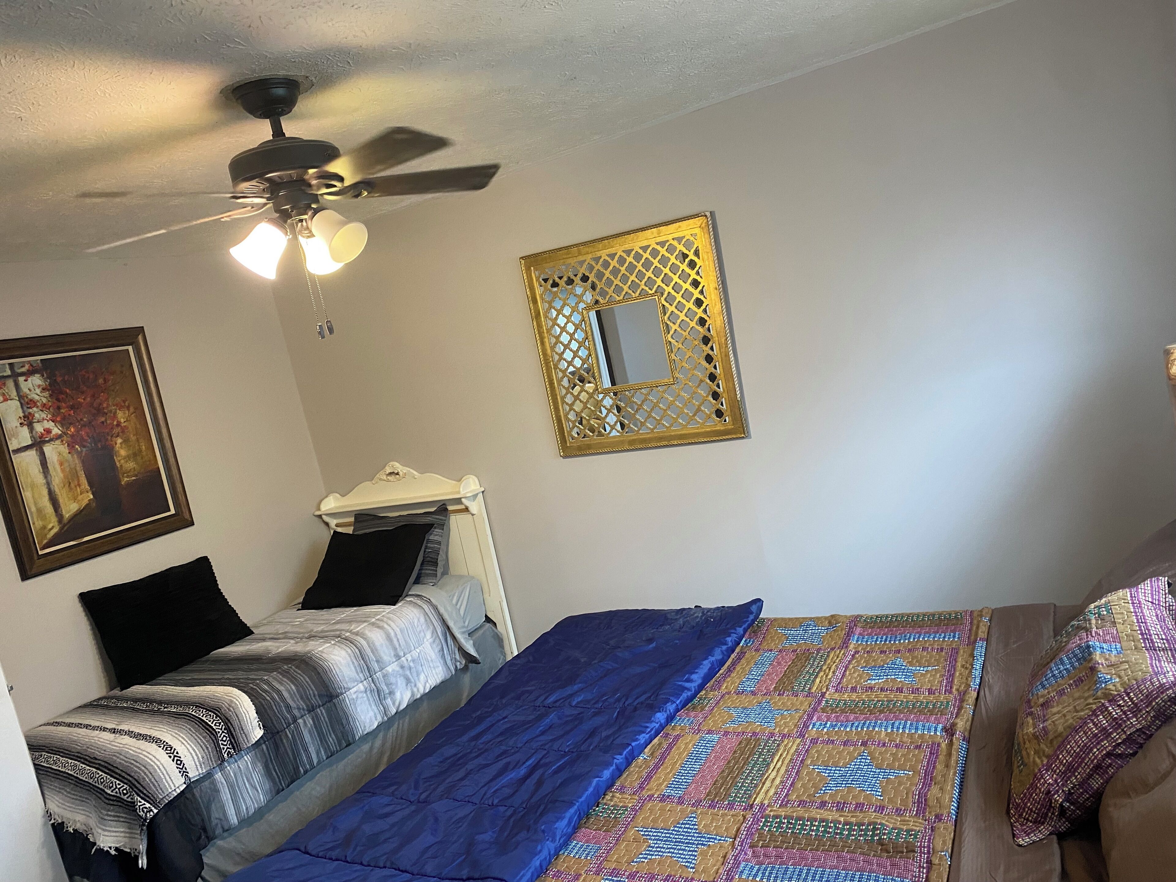 Un dormitorio con cama, ventilador de techo, espejo en la pared y un cuadro.
