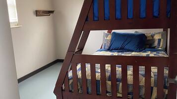 A bunk bed with a wooden frame and a blue and yellow quilt. There is a small shelf on the wall and a window with blinds.