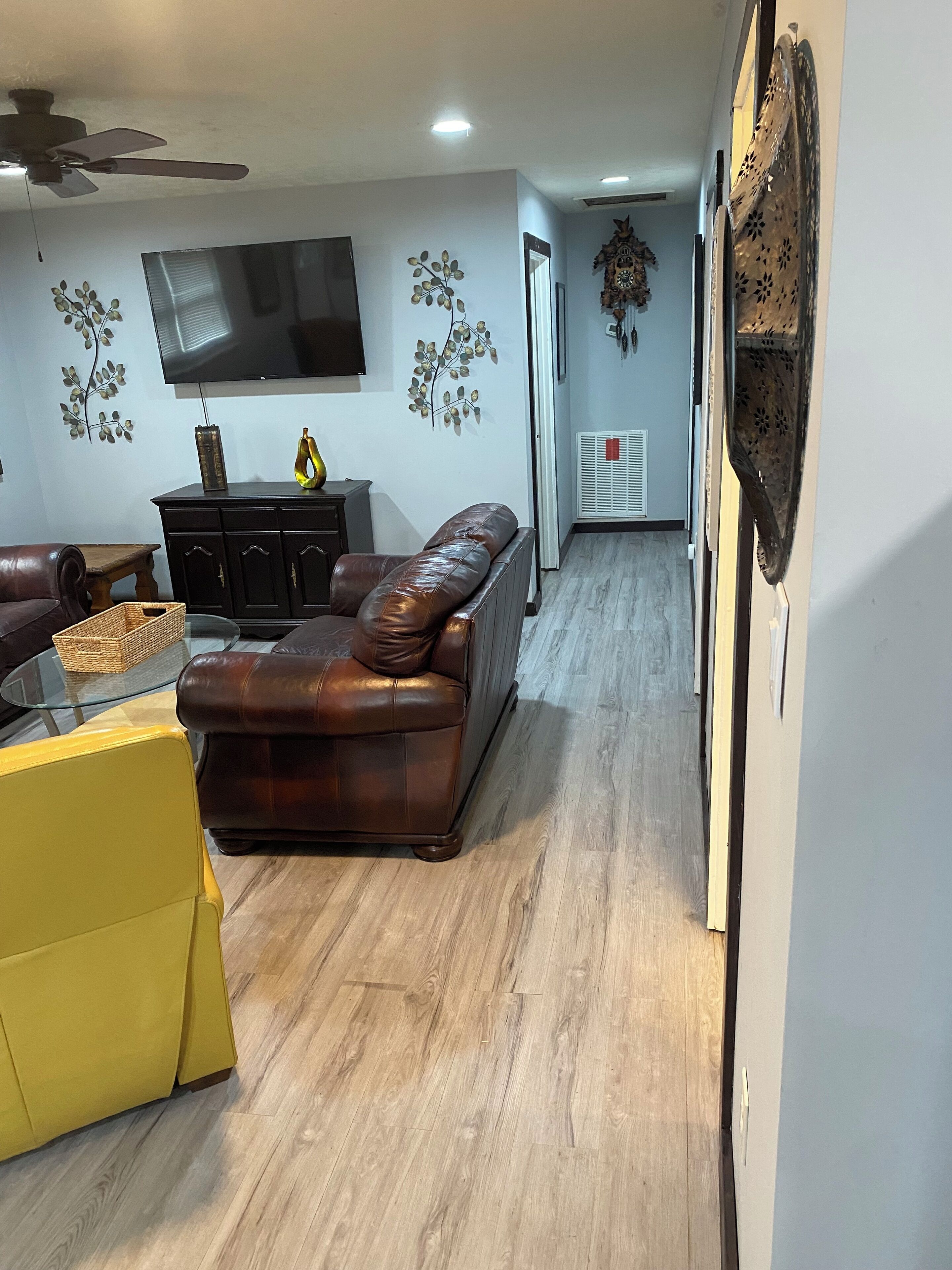 Ein Wohnzimmer mit braunem Ledersofa, einem gelben Sessel, einem Glastisch und einem an der Wand montierten Flachbildfernseher.