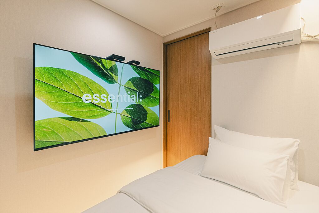 A modern hotel room with a large flat-screen TV mounted on the wall displaying a nature-themed wallpaper, a wooden door, and a white bed with pillows.