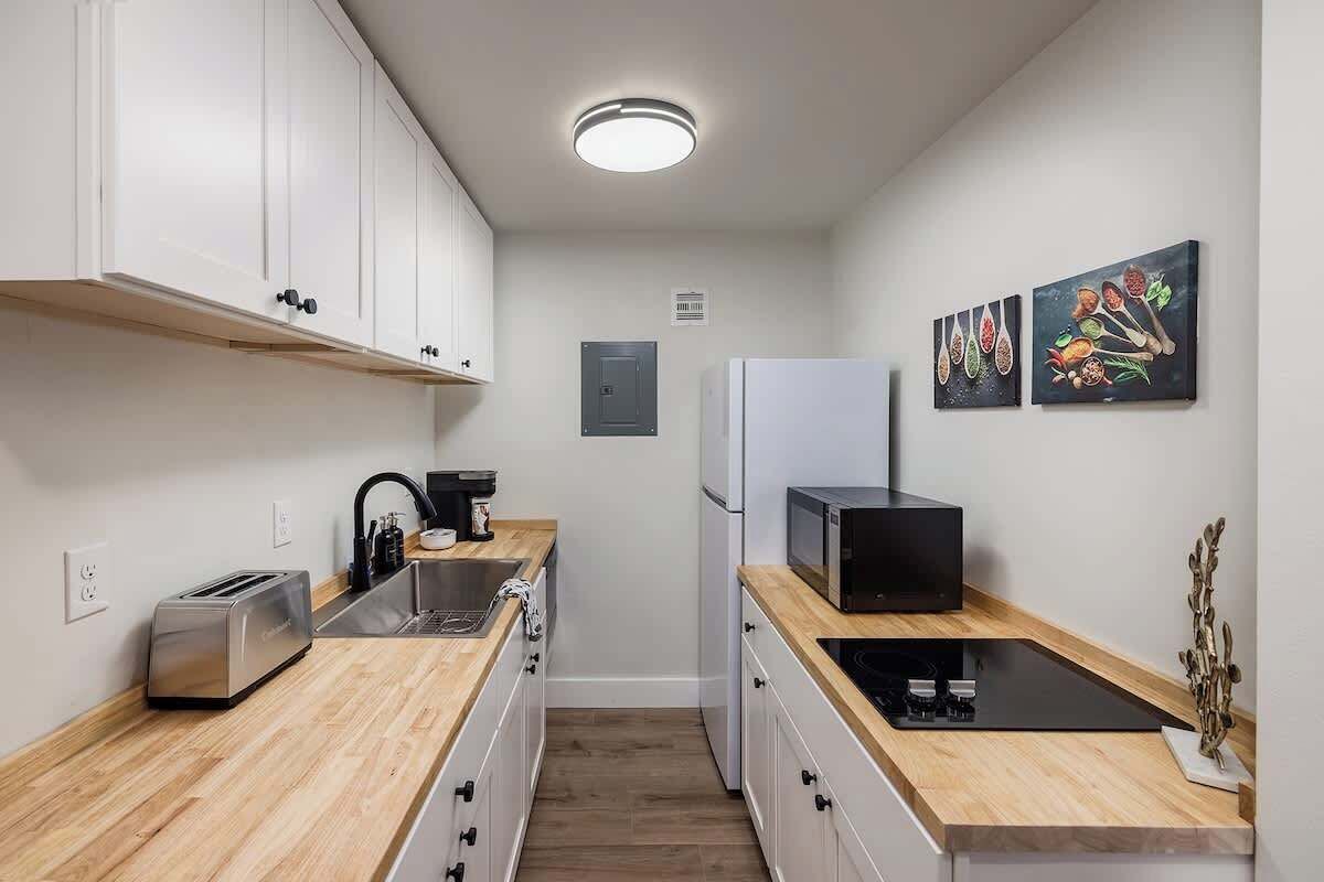 A compact kitchen with white cabinets, a toaster, a sink, a microwave, and a refrigerator. There are two wall-mounted artworks depicting fruits and vegetables.