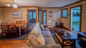 A spacious living room with a wooden dining table, a sofa, and a coffee table. There is a door leading to a balcony with a view of the outdoors. - The Guest House & Cottage at Seagate- Coos Bay, OR (Coos Bay)