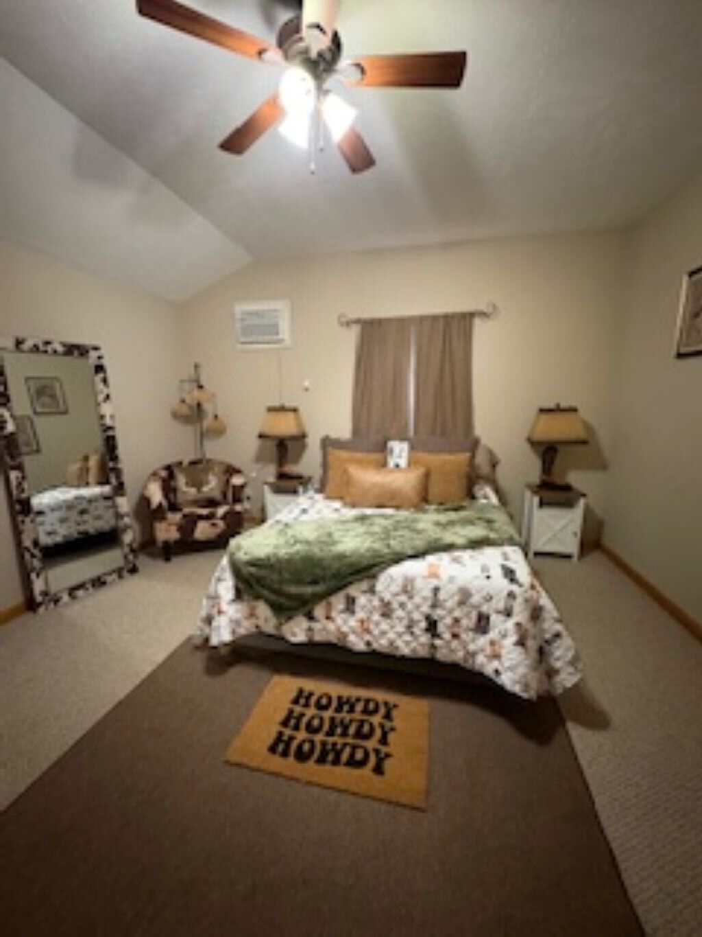 A bedroom with a bed, a ceiling fan, two bedside tables with lamps, a window with curtains, and a mirror with a patterned frame.