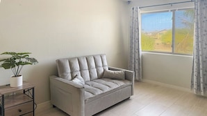A room with a sofa, a small wooden side table with drawers, a potted plant, and a window with curtains. - 3BR Casa Grande Home with Private Pool and Sofa Sleeper (Casa Grande)