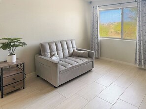 A room with a sofa, a small wooden side table with drawers, a potted plant, and a window with curtains. - 3BR Casa Grande Home with Private Pool and Sofa Sleeper (Casa Grande)