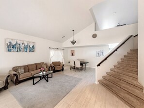Un área de estar amplia con sofá, mesa de centro y mesa de comedor. Una escalera conduce a un nivel superior.