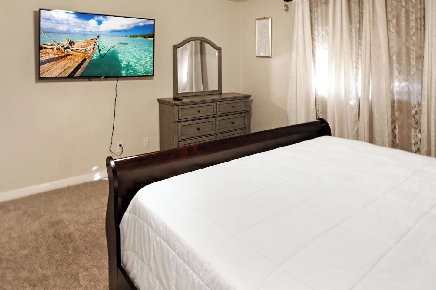 A bedroom with a bed, a dresser, a television mounted on the wall, and a window with curtains.
