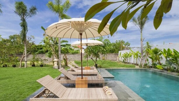 A poolside area with lounge chairs and umbrellas, surrounded by greenery and a clear sky. - Braya Living (Denpasar)