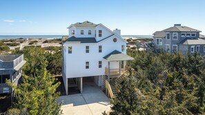 A two-story white house with a covered porch and a garage, surrounded by dense trees and a beach in the background.