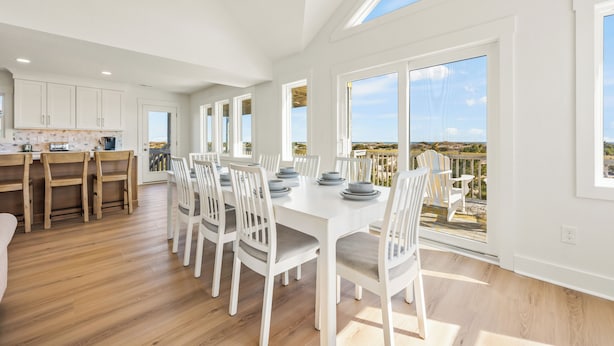 A spacious dining area with a long table, white chairs, and a view of the outdoors through large windows. - NEWLY RENOVATED! Oceanfront, Beach Access, Dog Friendly, Spacious Decks (Salvo)
